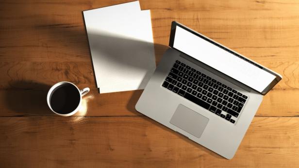Ein Laptop, ein Blatt Papier und eine Tasse Kaffee auf einem Holztisch.