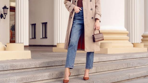 Auf dem Bild ist eine Frau zu sehen, die modisch gekleidet ist mit Jean, Trenchcoat und Tasche in der Hand.