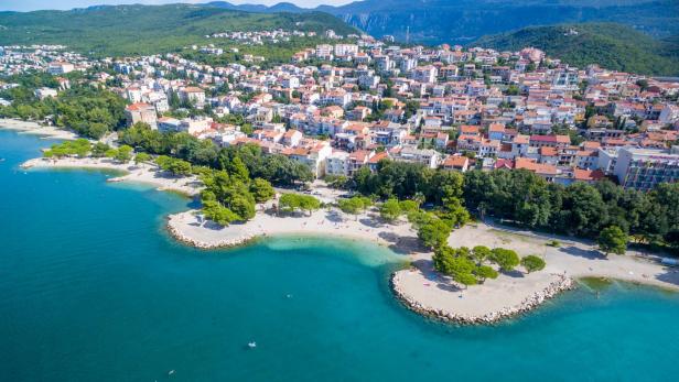 Luftaufnahme der Kvarner Bucht in Kroatien mit türkisblauem Meer, feinem Strand, grünen Pinienbäumen und malerischer Küstenstadt mit roten Ziegeldächern im Hintergrund.