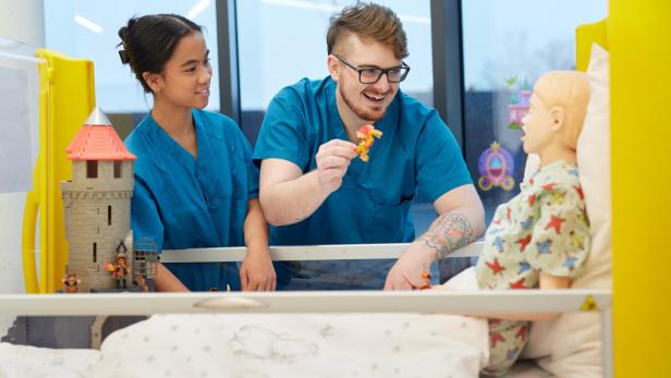 Zwei Pfleger:innen bespaßen eine Puppe in einem Krankenbett. 