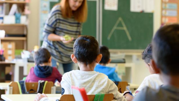Mehrere Kinder sitzen an Tischen in einem Klassenzimmer, während eine Lehrerin vorne an der Tafel steht.
