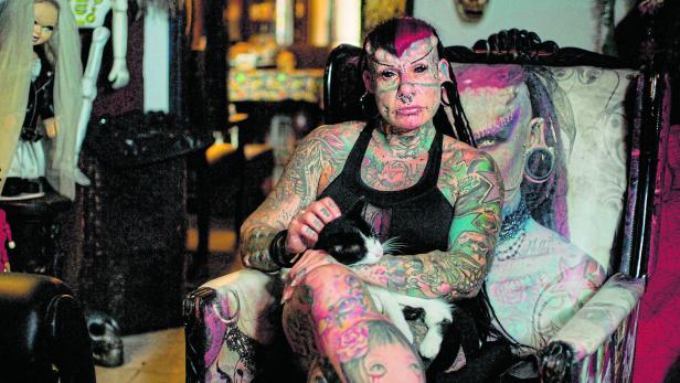 Eine stark tätowierte und modifizierte Frau sitzt mit einer Katze auf einem dekorierten Stuhl.