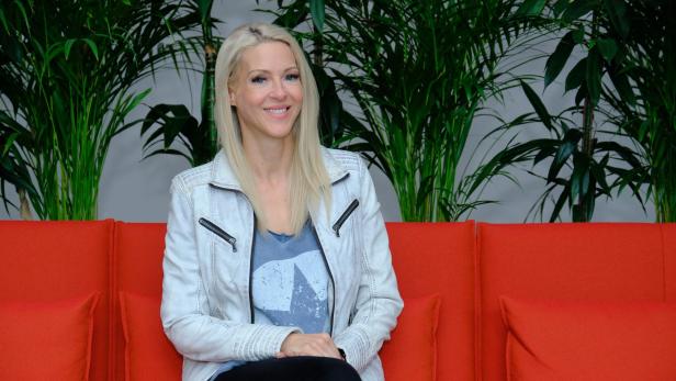 Simone Lugner sitzt auf einem roten Sofa vor grünen Pflanzen.