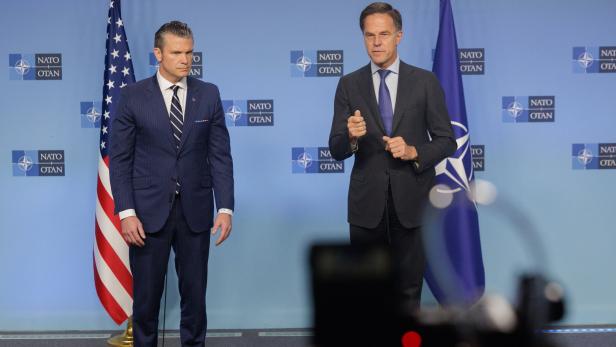 Zwei Männer in Anzügen stehen vor Flaggen der NATO und der USA.