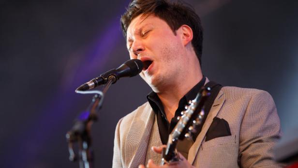 Ein Mann mit Sakko singt in ein Mikrofon und spielt Gitarre.