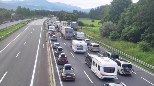 Stau auf der Autobahn mit vielen Autos und Lkw vor grüner Bergkulisse.