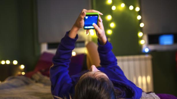 Jugendliche mit Smartphone auf einem Bett. KI-Apps sollen ihr Leben erleichtern.
