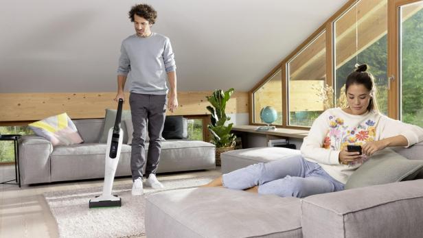 Eine Frau sitzt auf der Couch im Wohnzimmer und tippt auf ihrem Handy, während der Mann den Teppich staubsaugt.