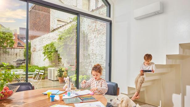 Zwei Kinder spielen in einem Wohnzimmer. Dahinter ist eine Klimaanlage zu sehen.