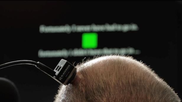 Ein Mann trägt ein schwarzes Neuroimplantat. Das Gerät ist direkt am Kopf befestigt. Im Hintergrund ist unscharf ein weißer Text auf einem Bildschirm zu sehen.