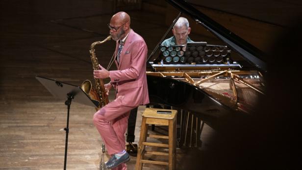 Ein Mann im rosa Anzug spielt Saxophon, während ein anderer Mann Klavier spielt.