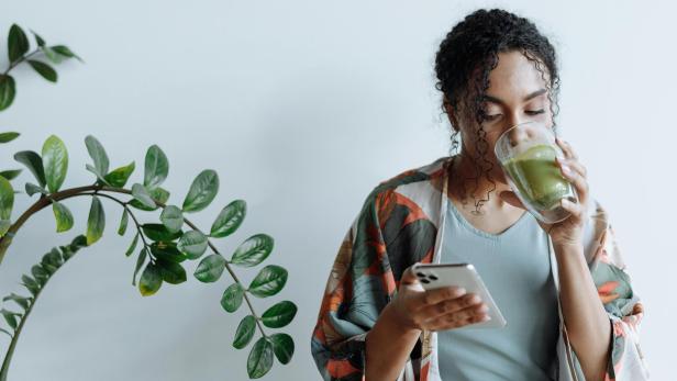 Person trinkt, während sie auf ihr Smartphone blickt, das sie in der Hand hält.