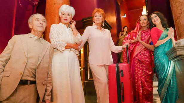 Eine Gruppe älterer Menschen mit Gepäck wird von zwei Frauen in Saris begrüßt.