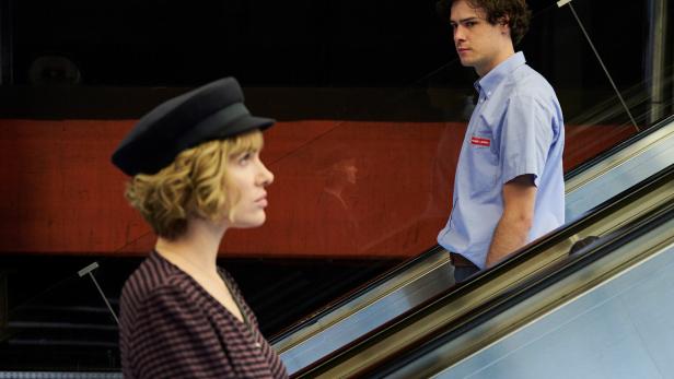 Eine Frau mit Hut und ein Mann mit Uniform stehen auf einer Rolltreppe.