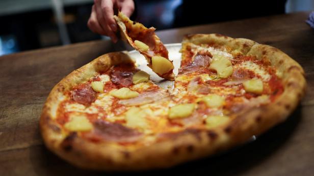 Eine Hand nimmt ein Stück Pizza mit Ananas und Schinken vom Teller.