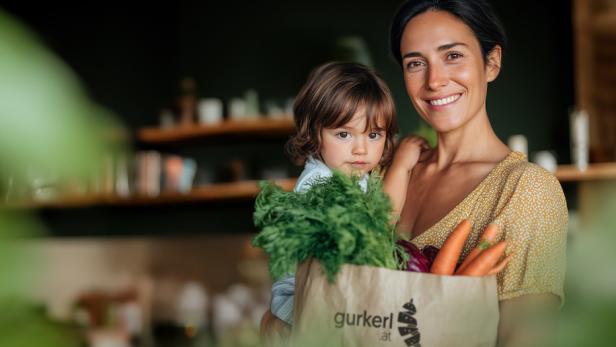 Eine Frau hat ihr Kind auf ihrem Arm und freut sich über ihre Einkaufstüte von gurkerl. 