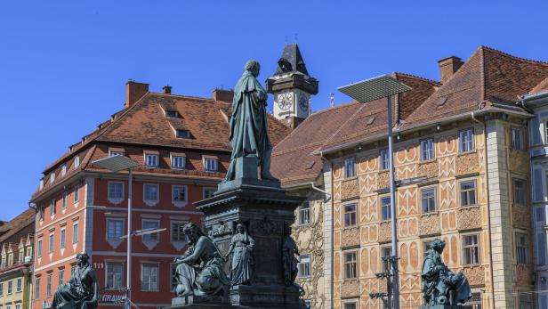 Eine Statue steht vor historischen Gebäuden in Graz, darunter der Grazer Uhrturm.