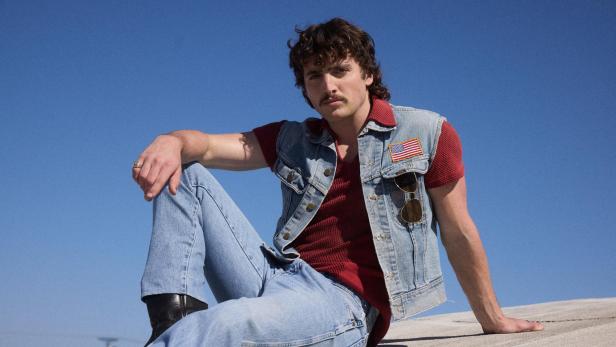 Benson Boone sitzt in Jeans und Jeansweste mit US-Flagge vor blauem Himmel.