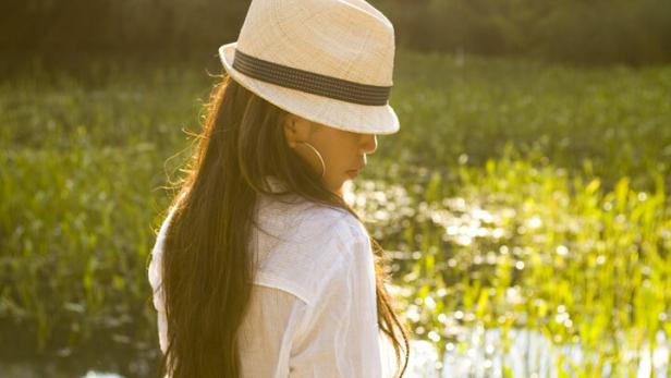 Eine Frau mit Hut steht an einem See im Gegenlicht.