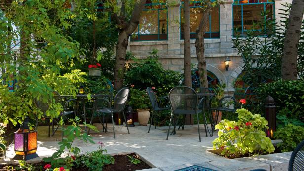 Schattiger Gartenhof mit Metallstühlen, Tischen, Laternen und blühenden Pflanzen vor einer Steinfassade mit bunten Fenstern.