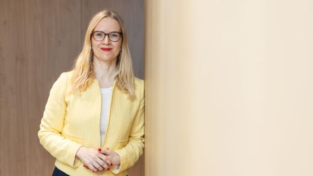Eva Aschauer steht angelehnt an einer Wand und trägt einen gelben Blazer.