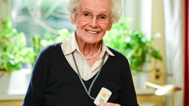 Eine ältere Dame mit einem mobilen Notrufgerät lächelt in die Kamera.