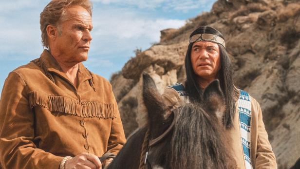 Zwei Männer in Westernkleidung reiten durch eine felsige Landschaft.