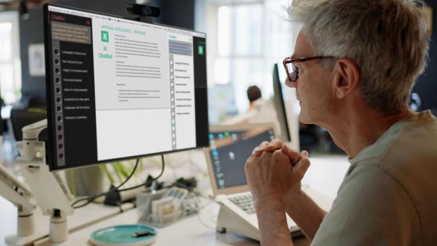 Ein älterer Mann mit Brille betrachtet einen KI-Chatbot auf einem Bildschirm im Büro.