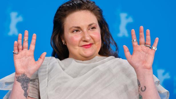 Lena Dunham posiert vor einem blauen Hintergrund und zeigt ihre Tattoos und Ringe.