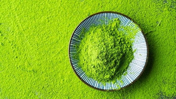 Eine kleine Schale mit leuchtend grünem Matcha-Pulver steht auf einer Fläche, die ebenfalls mit Pulver bedeckt ist.
