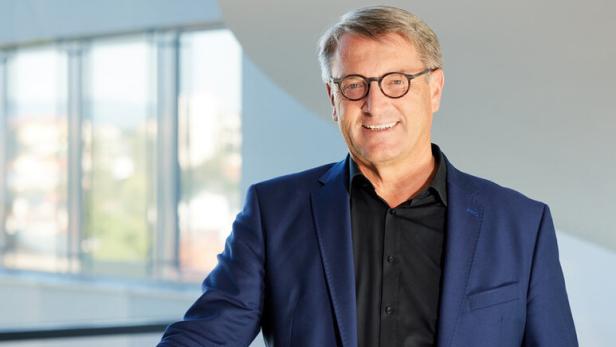 Ein lächelnder Mann mit Brille im blauen Sakko steht an einem Geländer in einem hellen Innenraum.
