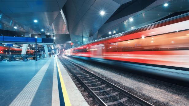 Ein roter Zug fährt mit hoher Geschwindigkeit durch einen modernen, beleuchteten Bahnhof.