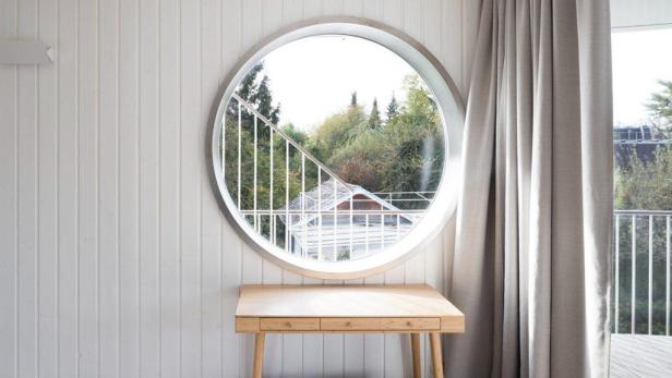 Ein runder Fensterblick auf ein Haus und Bäume über einem hellen Holzschreibtisch.