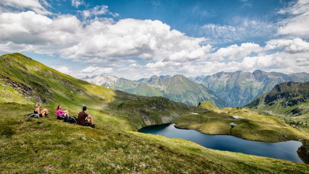 Drei Wanderer ruhen sich auf einem Hügel mit Blick auf einen Bergsee aus.