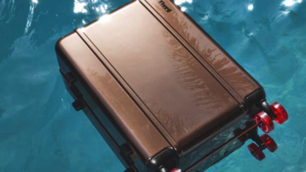Ein bronzefarbener Rollkoffer treibt auf dem türkisfarbenen Wasser.
