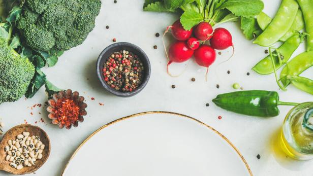 Frisches Gemüse, Getreide und Kräuter um einen leeren, weißen Teller herum.