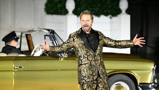 Ein Mann in einem auffälligen, gold-schwarz gemusterten Anzug gestikuliert vor einem goldenen Oldtimer mit Chauffeur.