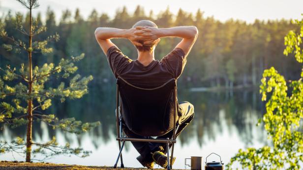 Man is sitting in a camping chair on the background of a forest lake on a beautiful summer evening.