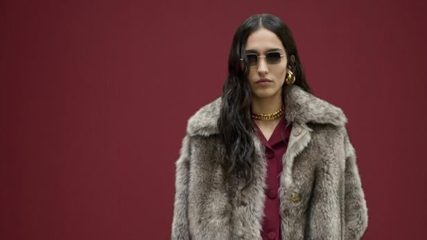 Eine Frau mit Sonnenbrille, Pelzmantel und Handtasche vor einem roten Hintergrund.