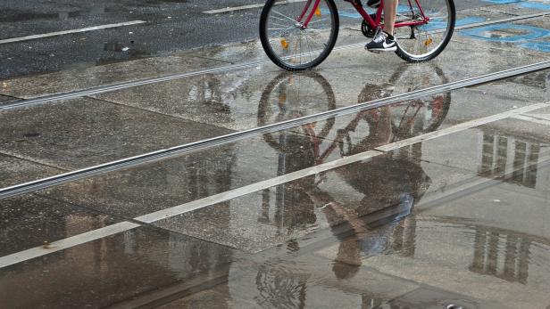 Ein Radfahrer fährt bei Regen über eine von Pfützen bedeckte Straße, in denen sich die Umgebung spiegelt.