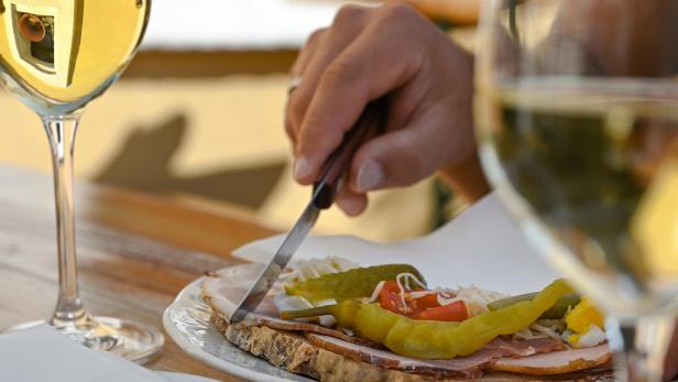 Eine Hand schneidet ein belegtes Brot mit Wurst, Käse, Tomate, Gurke und Peperoni, daneben stehen zwei Gläser Weißwein.