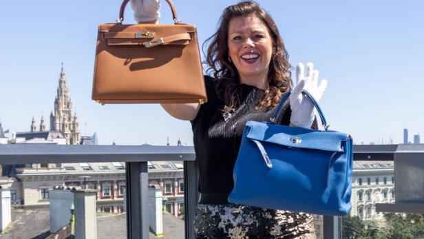 Eine Frau mit Handschuhen präsentiert zwei Handtaschen auf einer Dachterrasse in Wien.