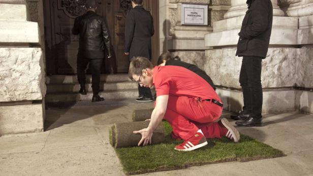 Ein Mann in roter Kleidung verlegt Rasenrollen vor einem Gebäude.