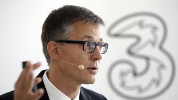 Ein Mann mit Brille und Mikrofon spricht vor einem Logo der Telefongesellschaft „3“.