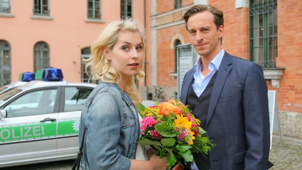 Eine Frau mit Blumenstrauß und ein Mann stehen vor einem Polizeiauto und einem Gebäude mit Backsteinfassade.