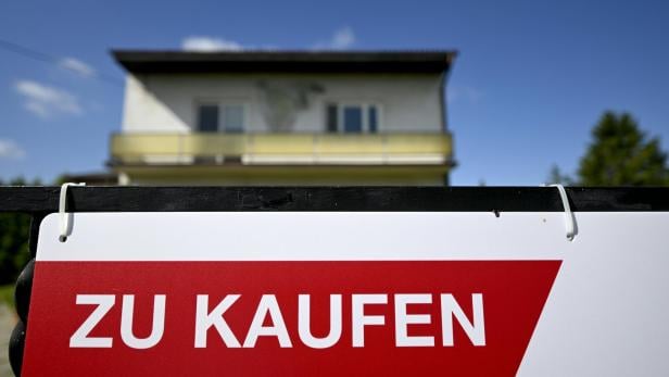 Ein zweistöckiges Haus mit Balkon steht im Hintergrund, davor ein Schild mit der Aufschrift „Zu kaufen“.
