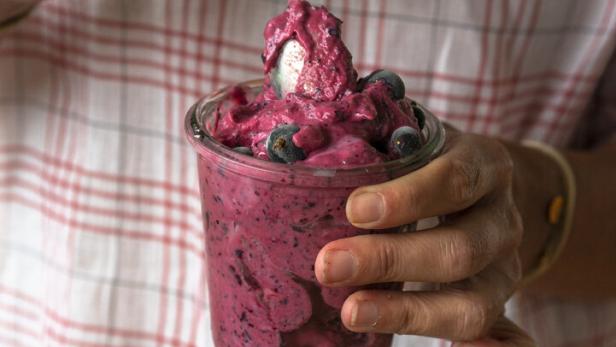 Eine Person hält ein Glas mit rosa Eis und Blaubeeren in der Hand.