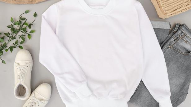 Ein weißer Pullover liegt schon aufgelegt auf dem Boden, daneben eine graue Jeans, weiße Sneaker und eine Korbtasche.
