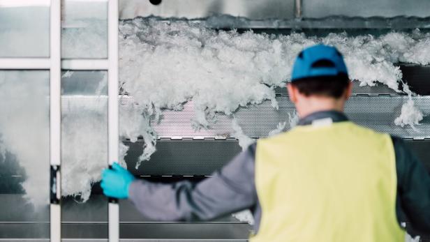 Arbeiter von Lenzing steht vor einer Fasertrocknungs-Maschine, aus der fluffige Fasern purzeln. Arbeiter von Lenzing steht vor einer Fasertrocknungs-Maschine, aus der fluffige Fasern purzeln.
