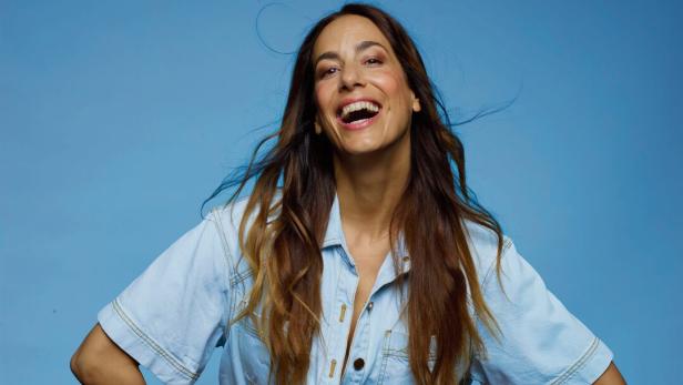 Eine lachende Frau in Jeanshemd und Shorts posiert vor blauem Hintergrund.